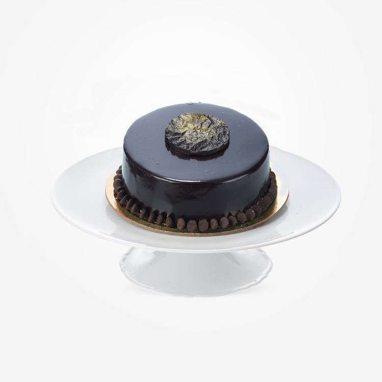 Glossy dark chocolate cake with gold leaf and chocolate beads on white stand.