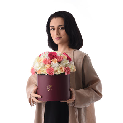 Woman holds burgundy box with pink, peach, white roses and carnations, gold logo, elegant beige robe.