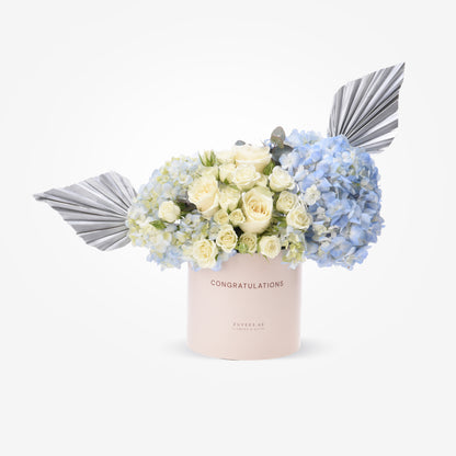 Pastel pink box with white roses and blue hydrangeas, accented by silver fan leaves.