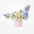 Pastel pink box with white roses and blue hydrangeas, accented by silver fan leaves.