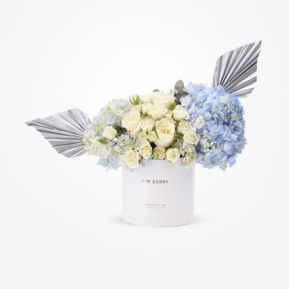 White box holds creamy roses and blue hydrangeas, accented with metallic silver wings.