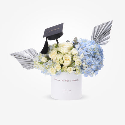 White roses and blue hydrangeas in sleek white box with graduation cap and silver wings.