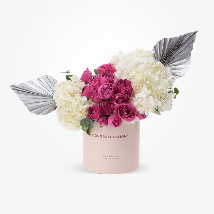 Pink vase with “CONGRATULATIONS” text, holding vibrant pink roses and white hydrangeas, accented by metallic silver fan leaves.