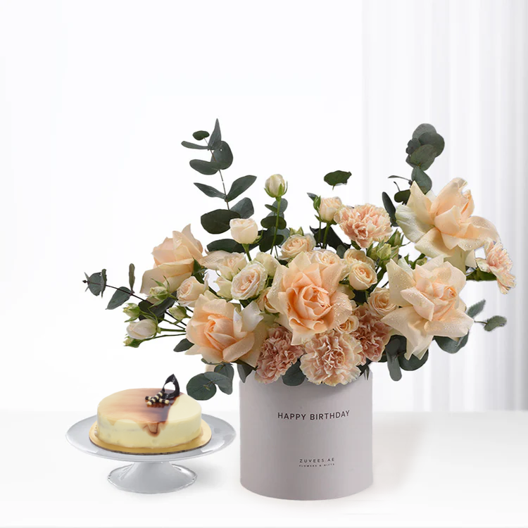 Soft peach roses and eucalyptus in gray “Happy Birthday” vase beside elegant cheesecake.