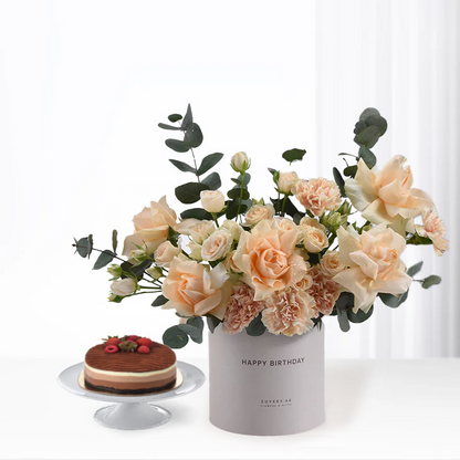 Soft peach roses and eucalyptus in gray "Happy Birthday" vase beside chocolate cake.