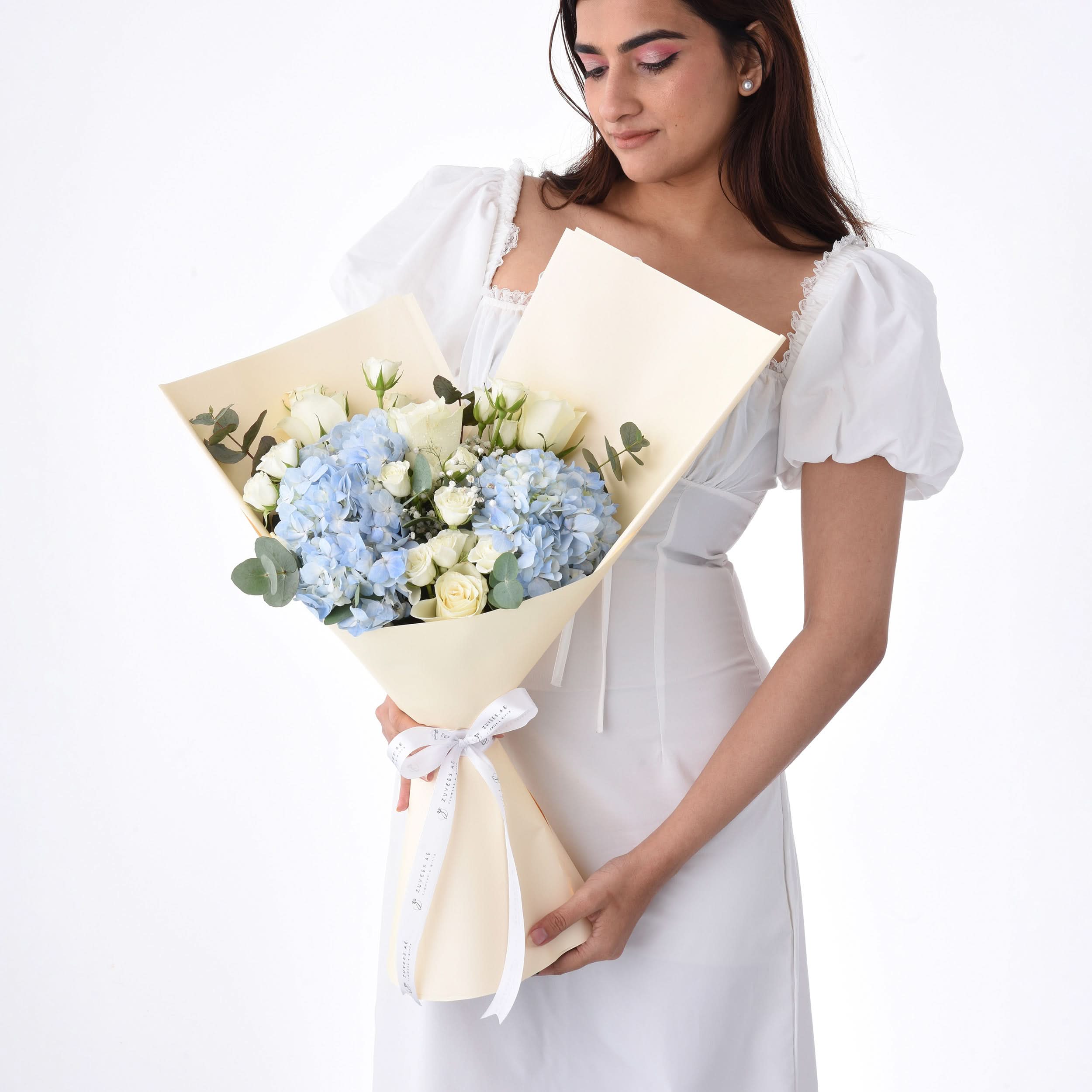 Woman in white dress holds elegant bouquet of blue hydrangeas and white roses, wrapped in cream paper with white ribbon.