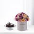 Purple, pink, and peach roses in grey "Happy Birthday" cylinder beside chocolate cake.