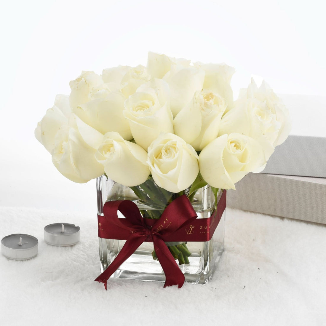 Elegant white roses in clear vase with burgundy ribbon, soft white backdrop, tea lights nearby.