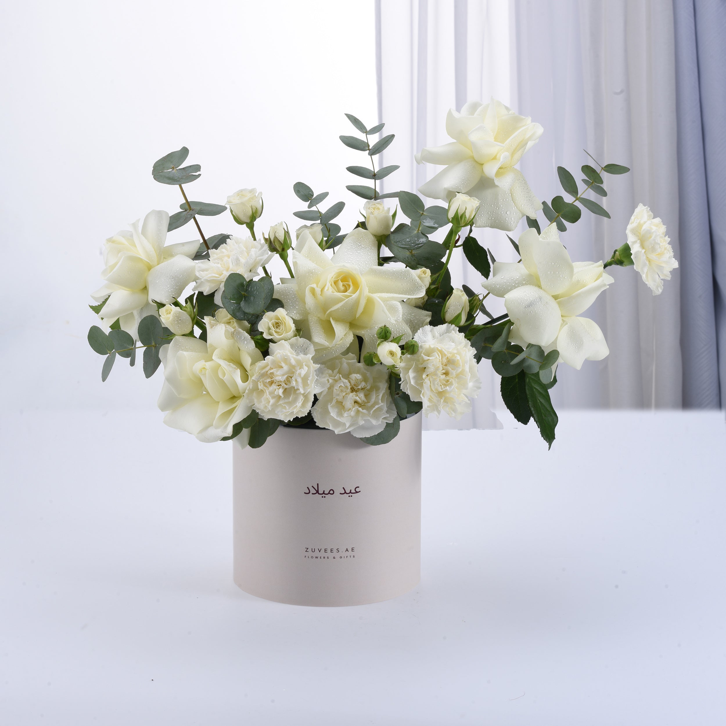 Elegant white rose and carnation bouquet in branded beige cylinder vase.