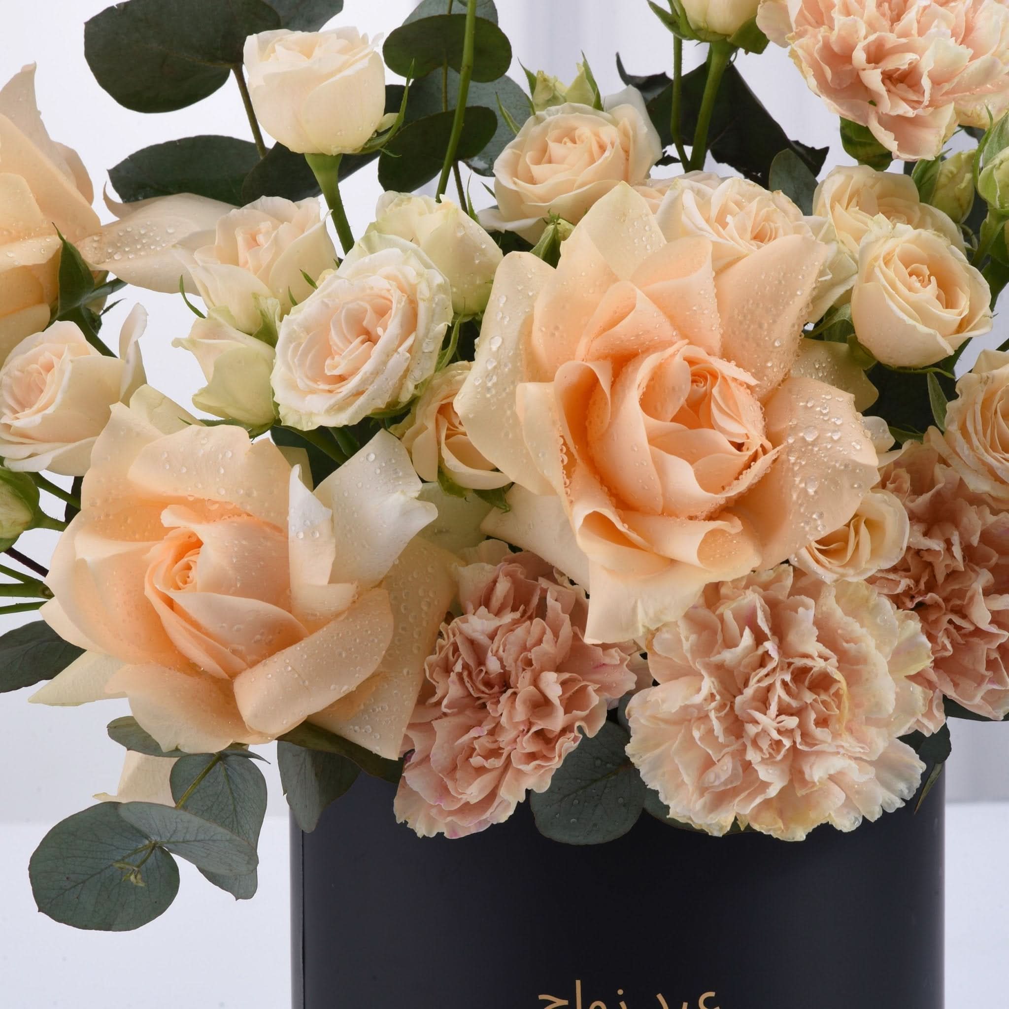 Soft peach roses and carnations glisten with dewdrops in sleek black vase.
