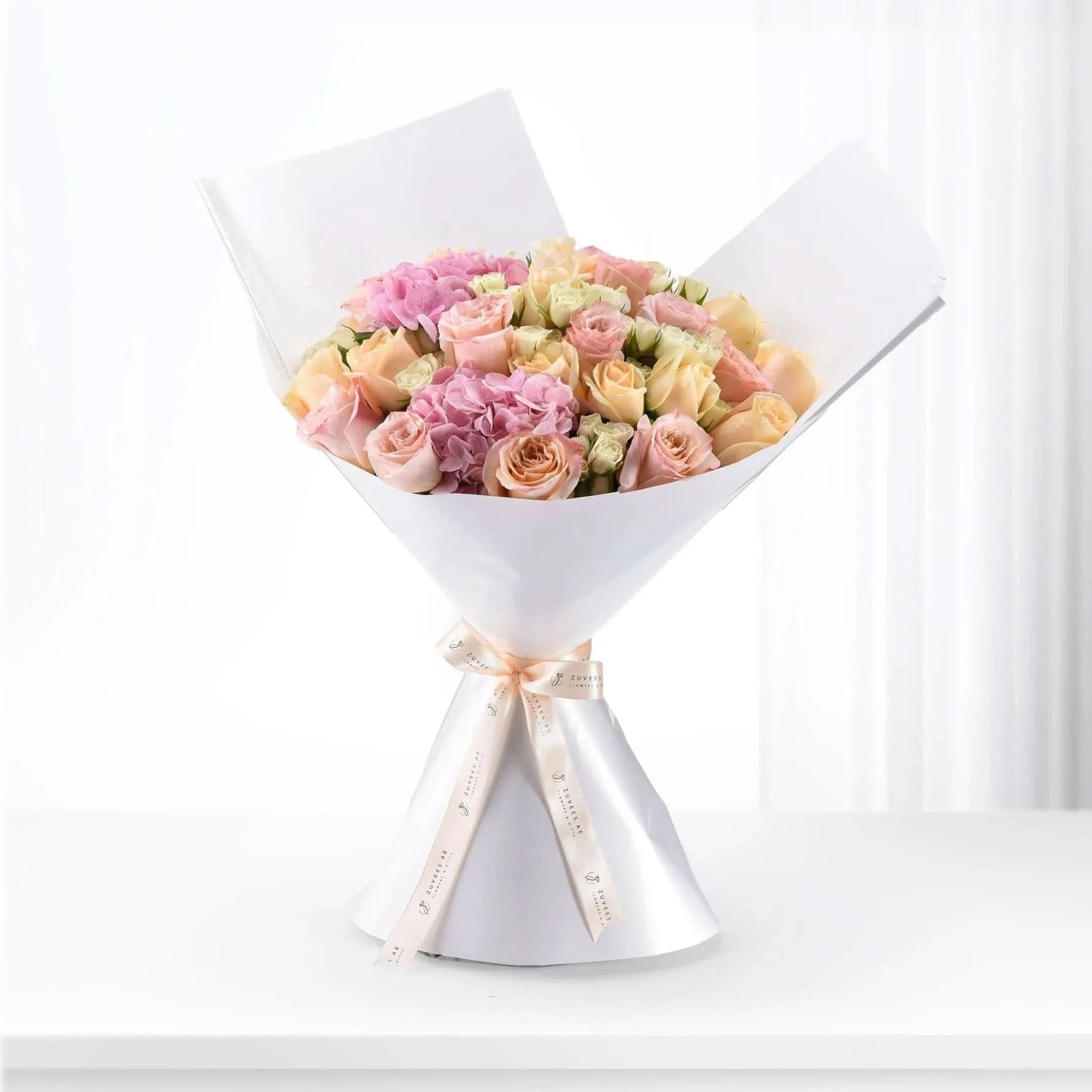 Soft pink and peach roses with hydrangeas, elegantly wrapped in white paper and ribbon.