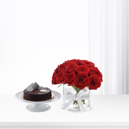 Vibrant red roses in glass vase beside rich chocolate cake on white pedestal.