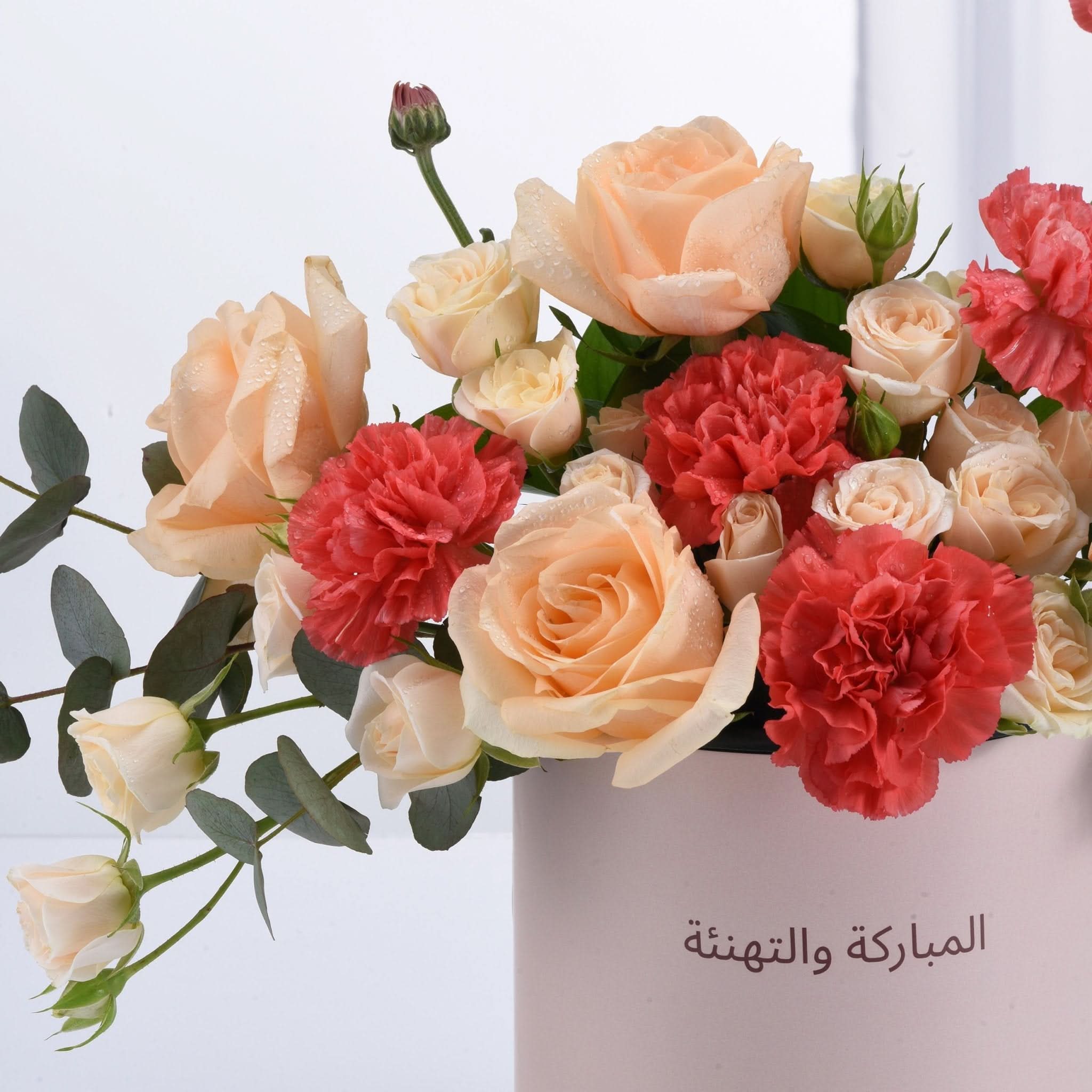 Soft peach roses and coral carnations in pink vase with Arabic "Congratulations" text.