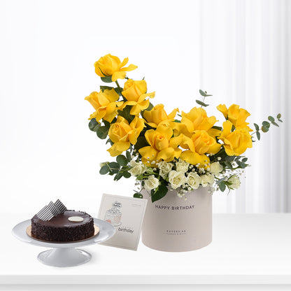Vibrant yellow roses and white blooms in beige "Happy Birthday" vase beside chocolate cake.