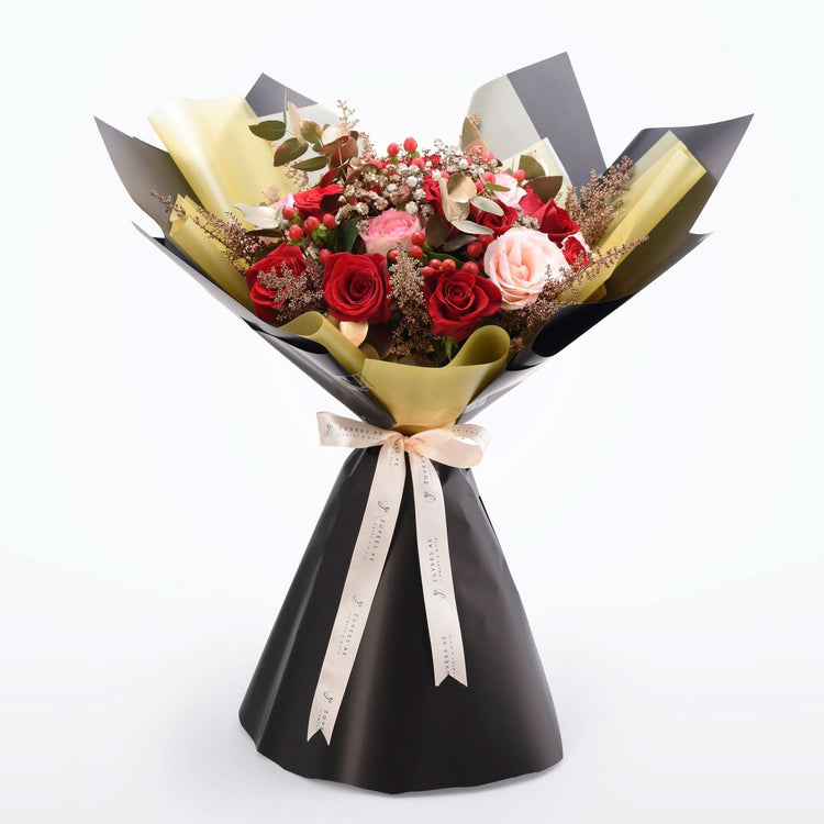 Elegant bouquet of red and pink roses, accented with berries and gold foil, wrapped in black and gold paper with branded ribbon.