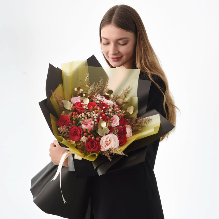 Woman holds lush red and pink rose bouquet wrapped in black and gold paper.