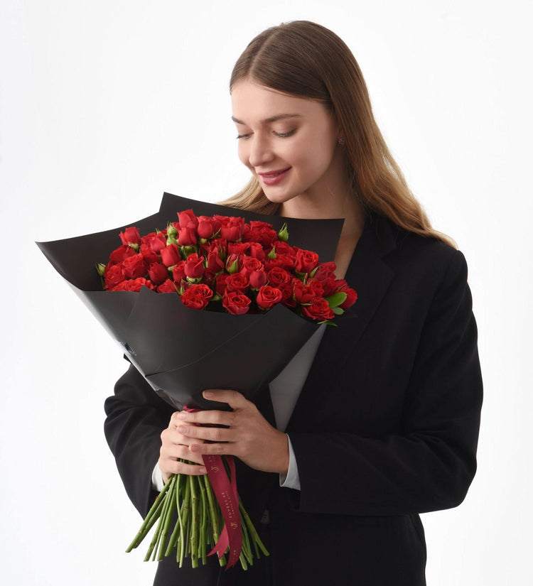 Woman in black suit gently holds vibrant red roses bouquet wrapped in sleek black paper.