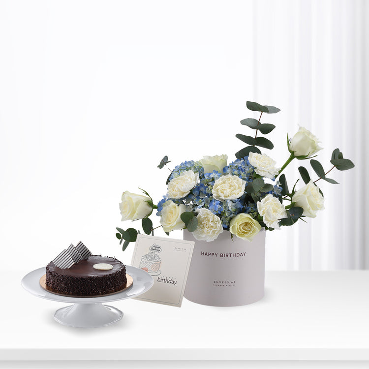 Elegant white roses and blue hydrangeas in gray box beside chocolate cake.