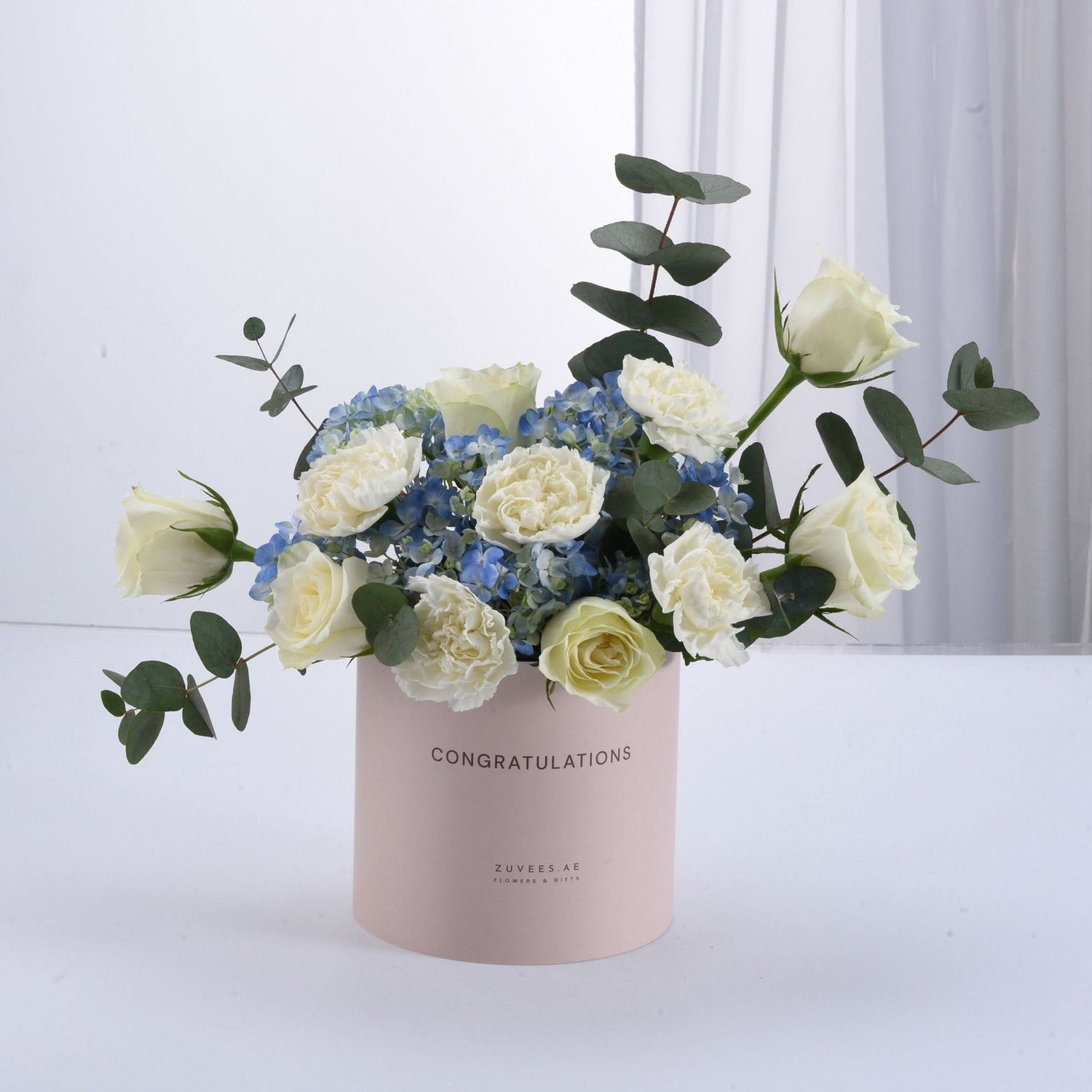Soft pink box holds white roses, blue hydrangeas, and eucalyptus—perfect for congratulations.