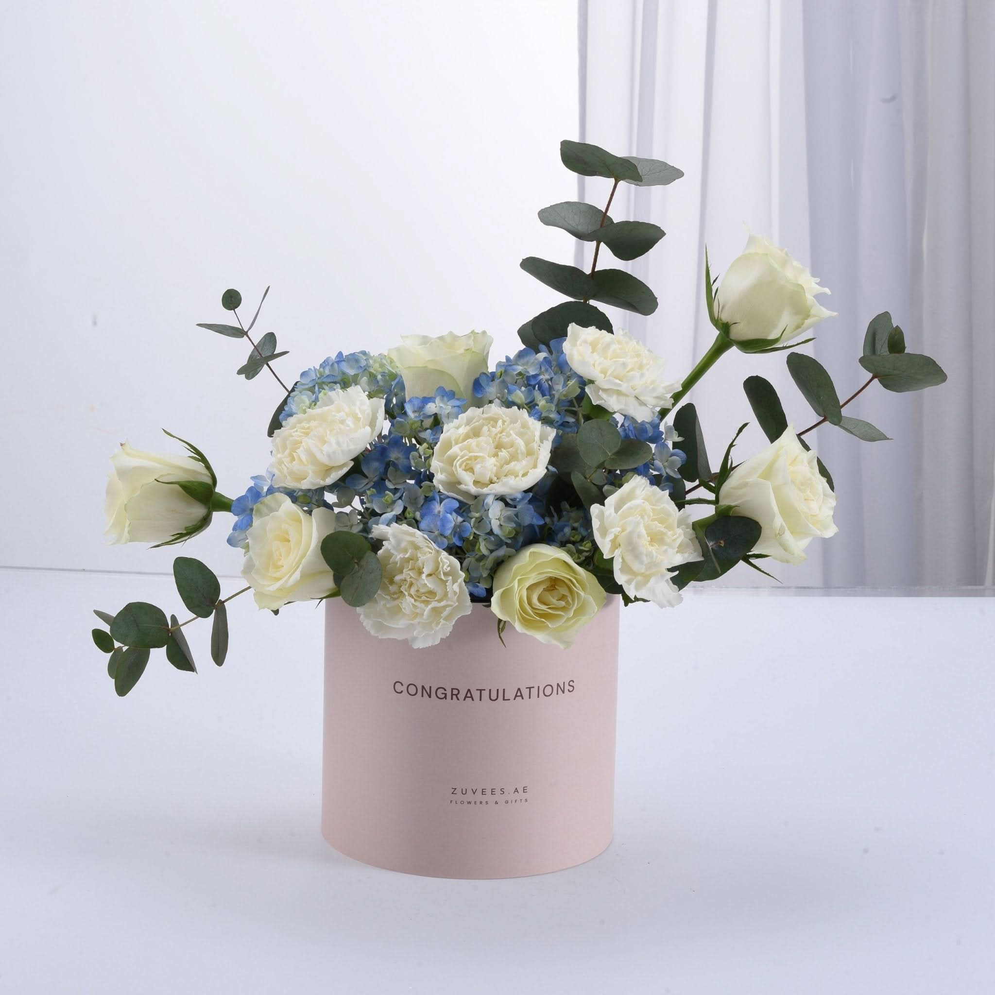 Soft pink box holds white roses, blue hydrangeas, and eucalyptus—perfect for congratulations.
