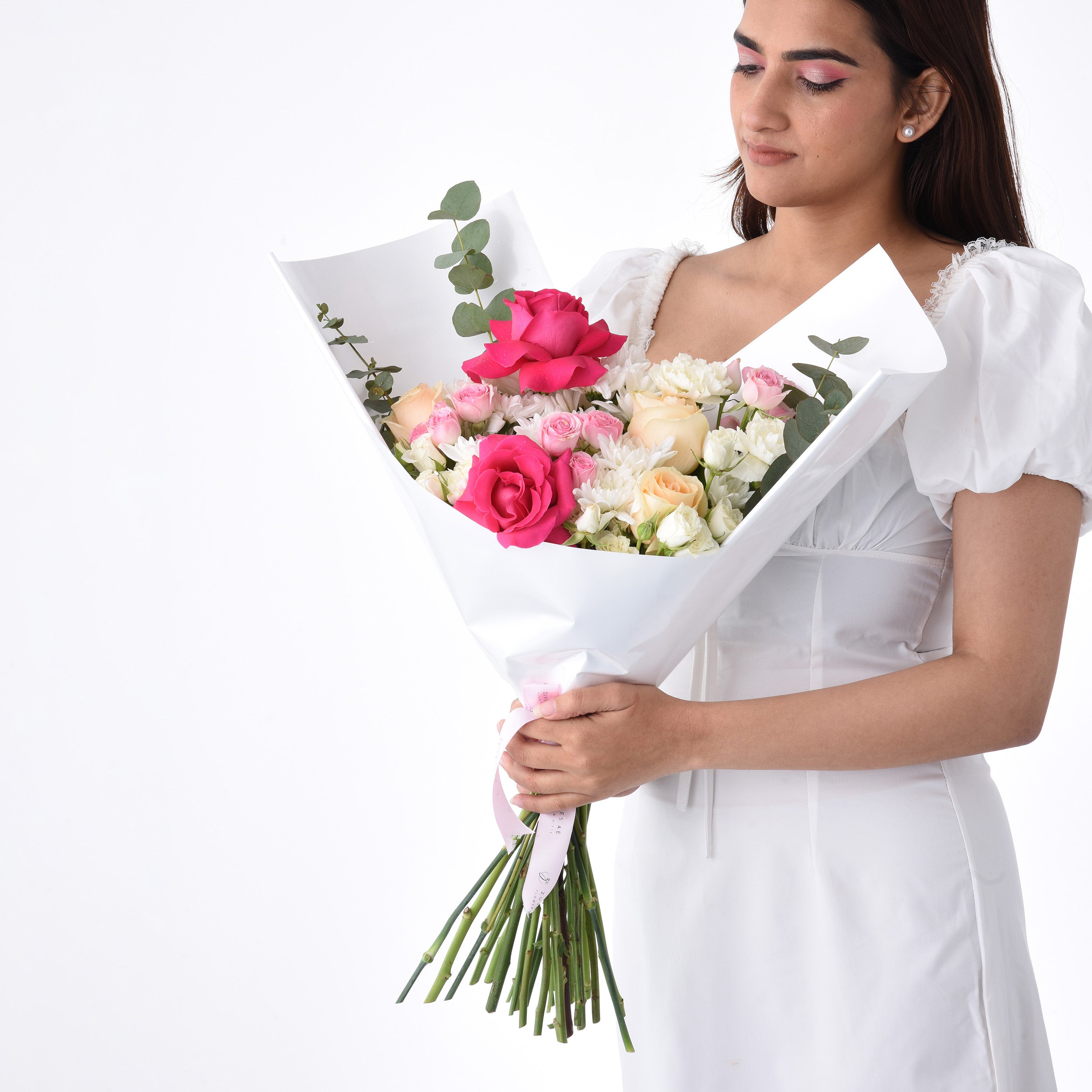 Woman in white dress holds vibrant pink, peach, and white rose bouquet wrapped in crisp white paper.