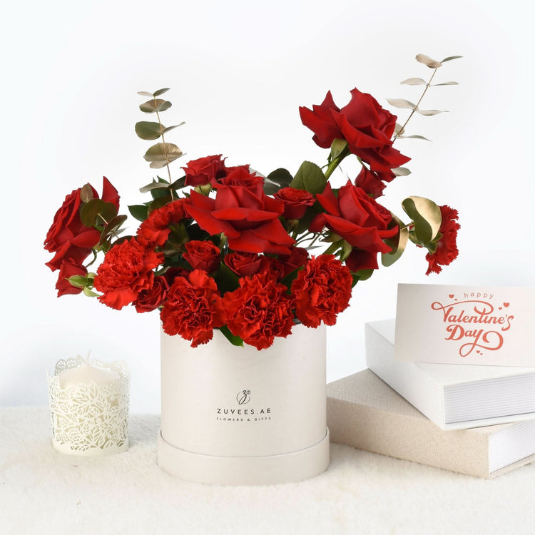 Vibrant red roses and carnations in white box beside Valentine’s card.
