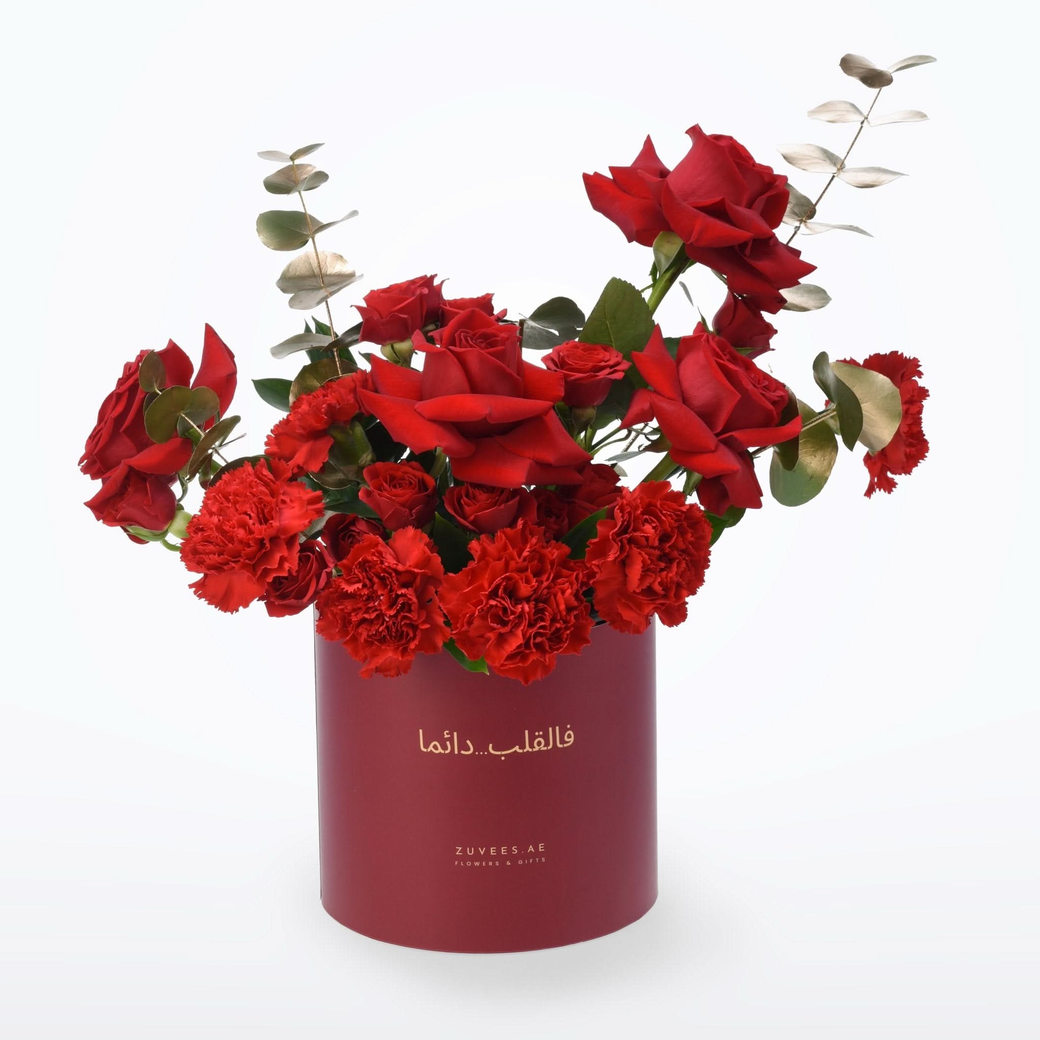 Vibrant red roses and carnations in deep burgundy cylindrical box with gold script.