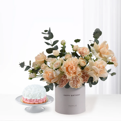 Soft peach roses and eucalyptus in gray vase beside pink-white cake.