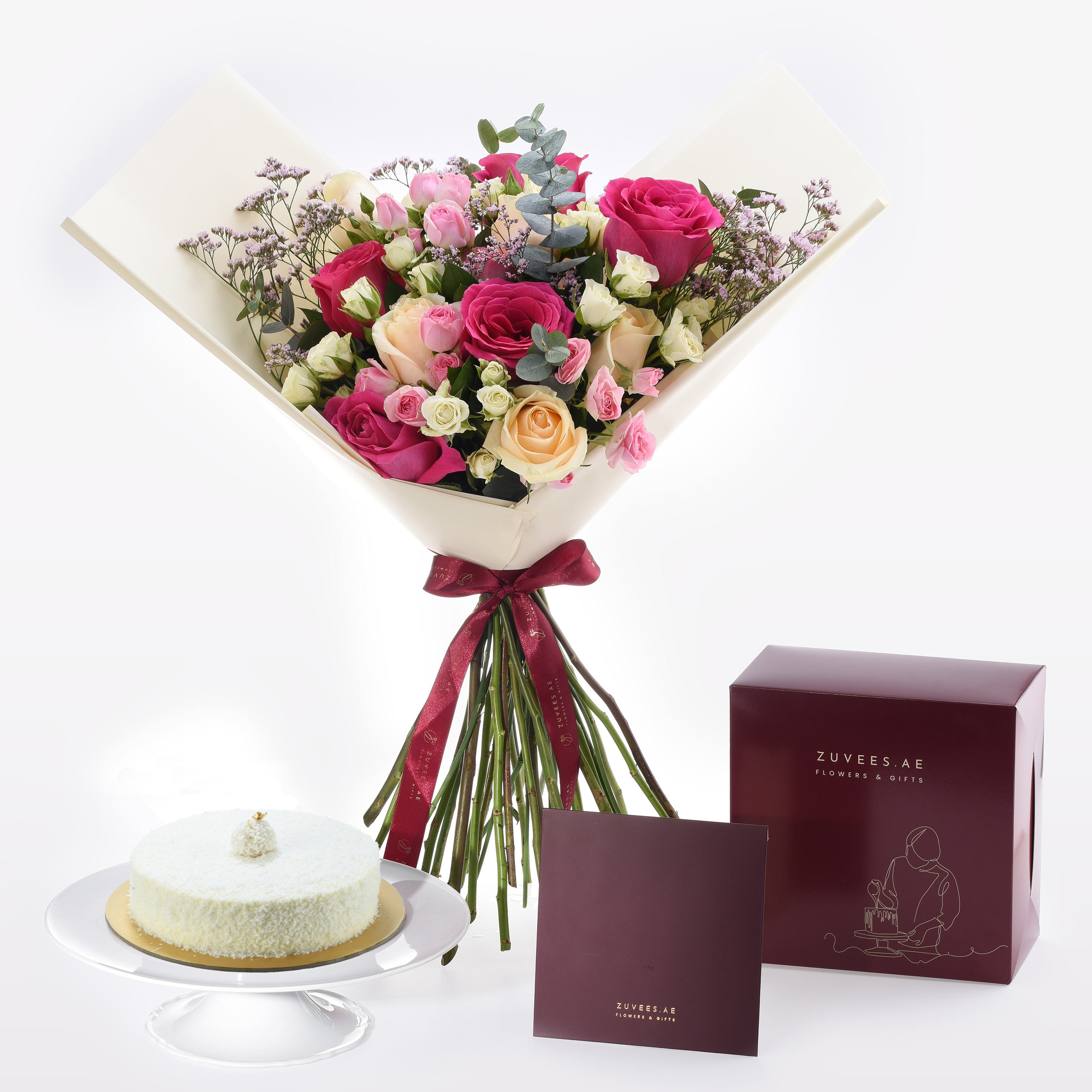 Vibrant pink, peach, and white roses with eucalyptus, wrapped in cream paper with burgundy ribbon, beside coconut cake and branded gift box.