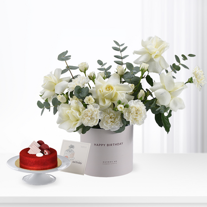 White roses and eucalyptus in gray box beside red velvet cake.