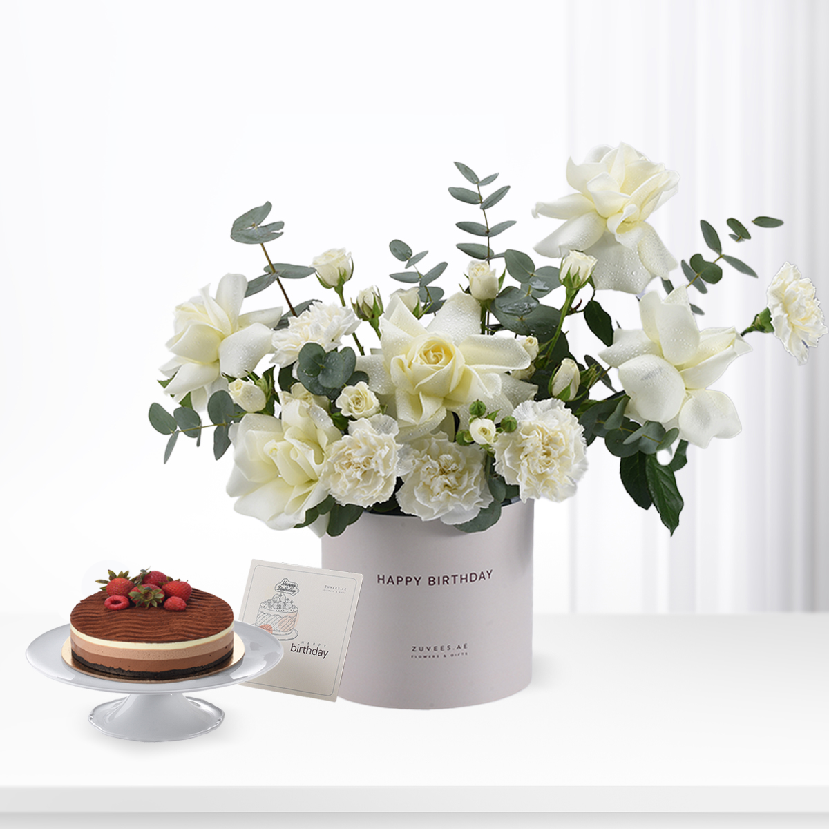 White roses and eucalyptus in branded box with birthday cake and card.