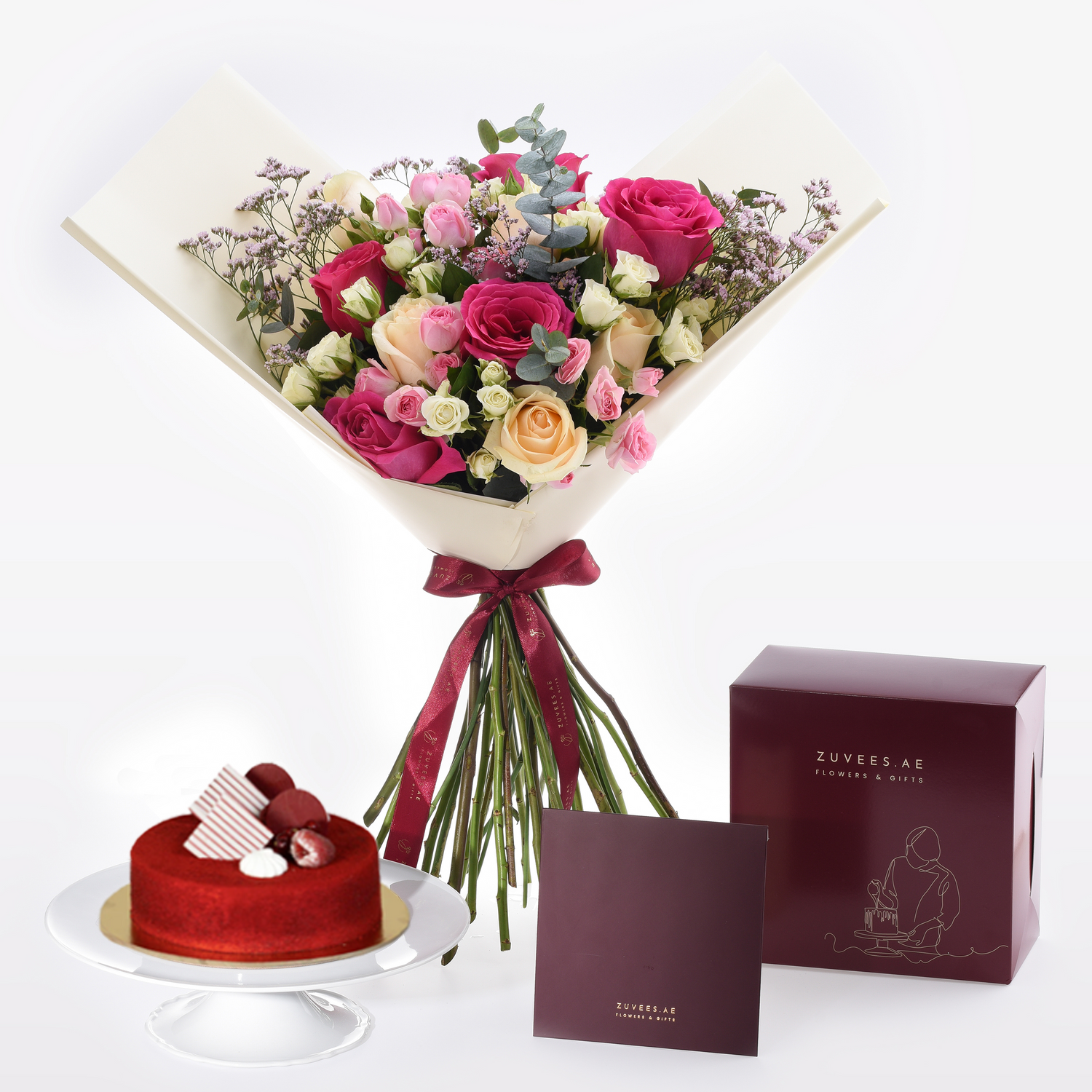 Vibrant pink rose bouquet with red ribbon, beside red velvet cake and branded gift boxes.