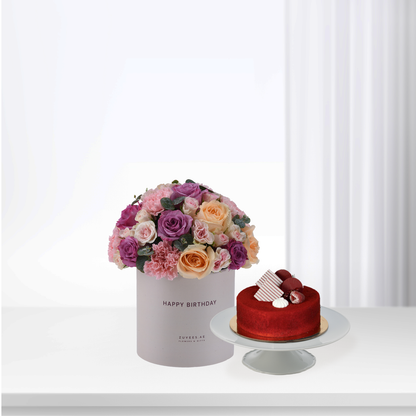 Elegant birthday bouquet with pink, purple, peach roses beside red velvet cake on white pedestal.