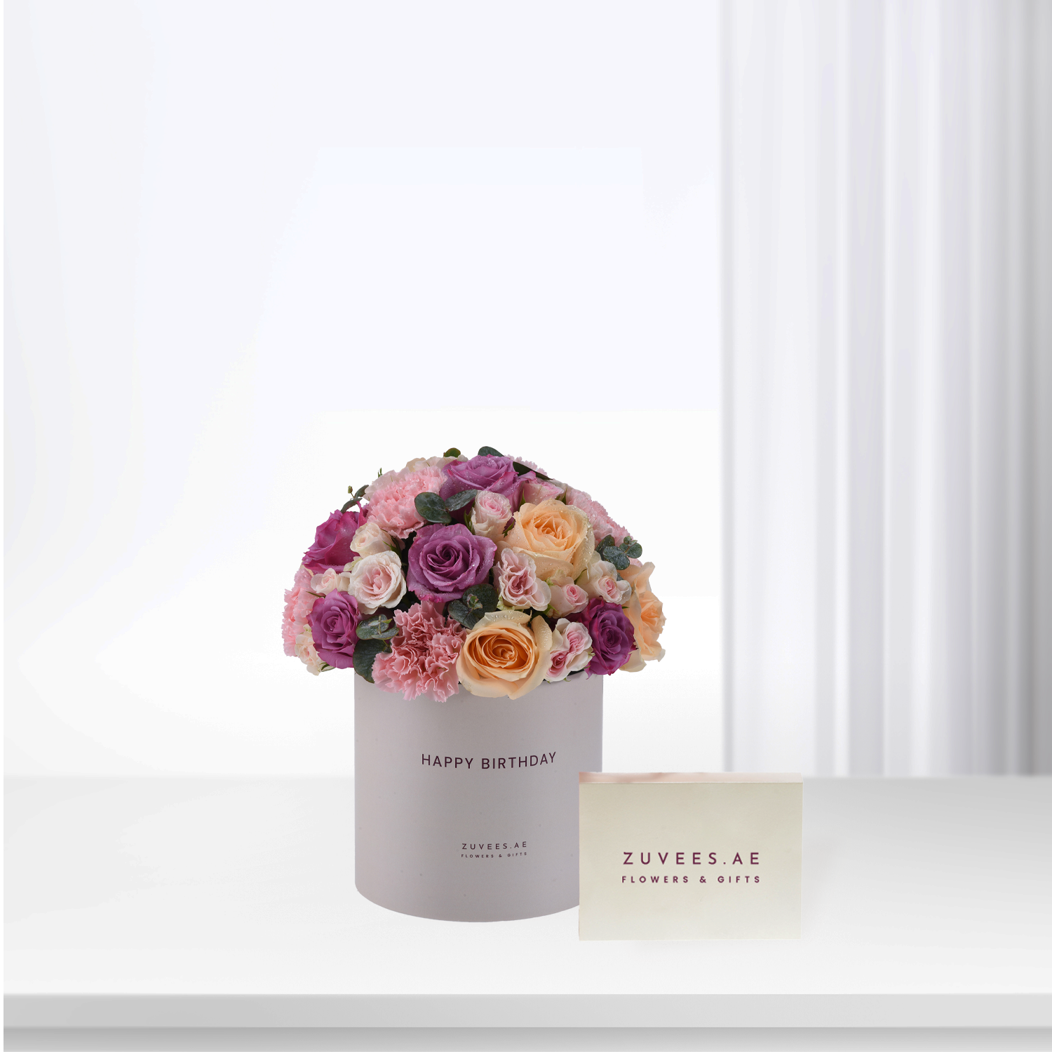Pastel pink, purple, and peach roses bloom in a branded birthday box beside matching card.