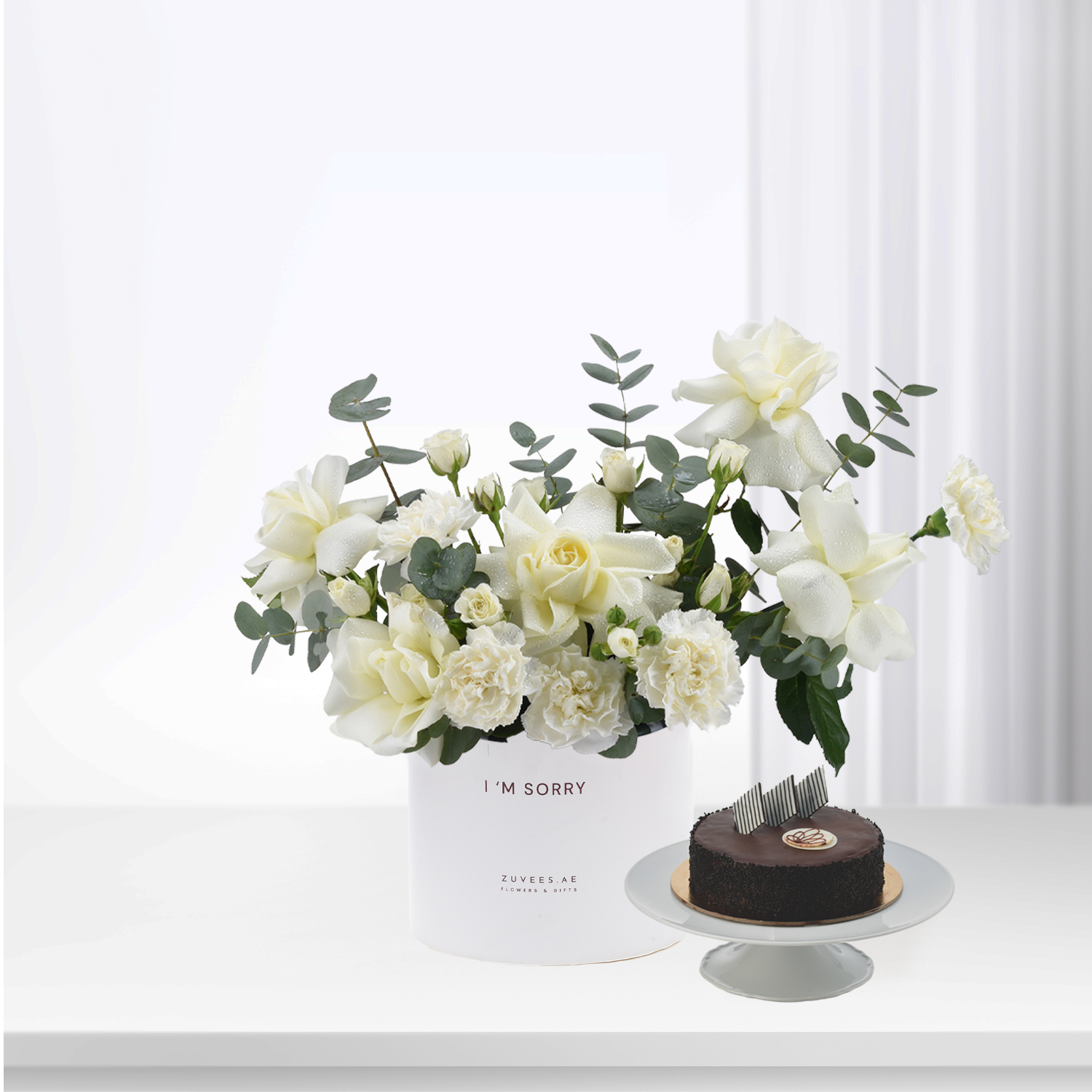 White roses and eucalyptus in minimalist vase beside chocolate cake.