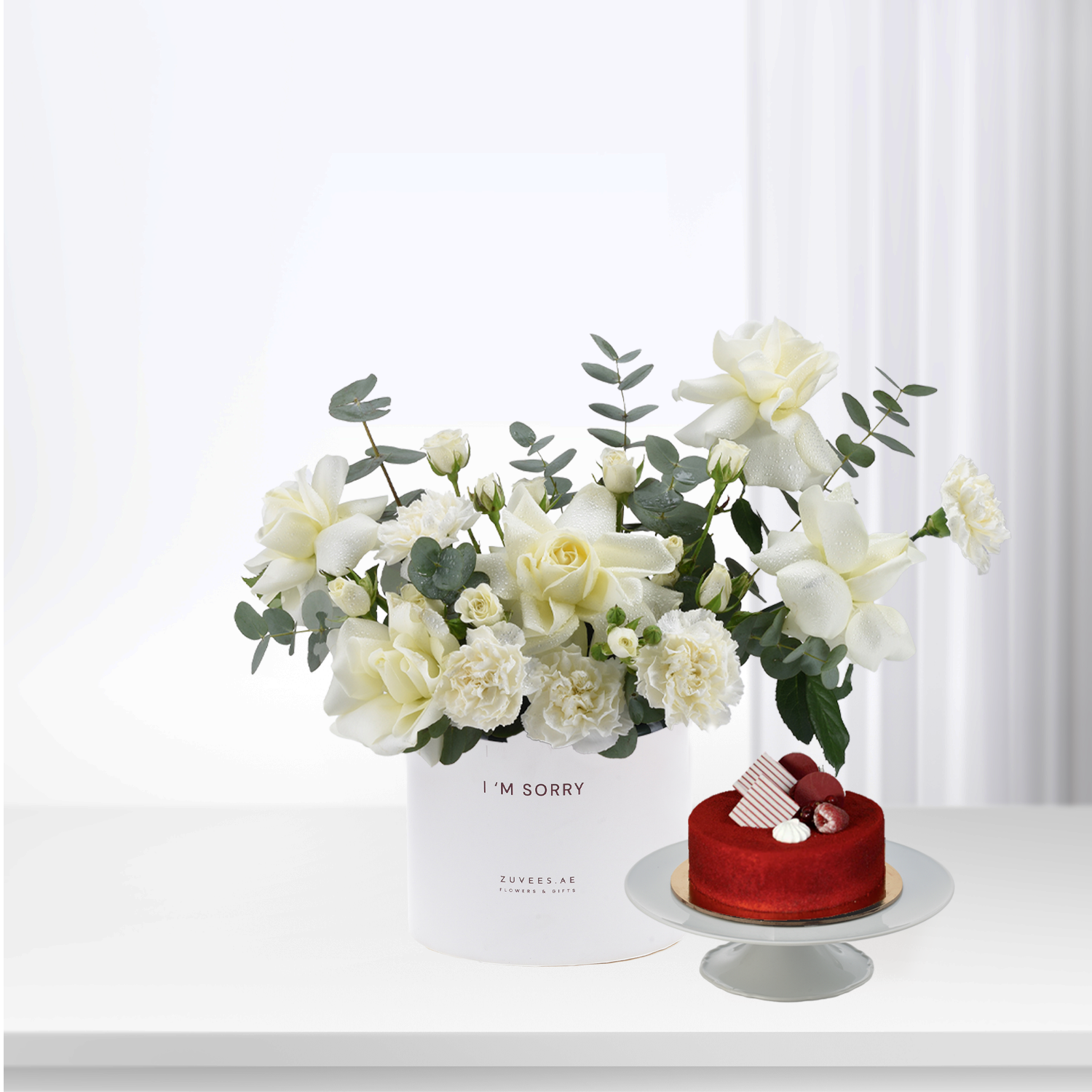 White roses and eucalyptus in minimalist box beside red velvet cake.