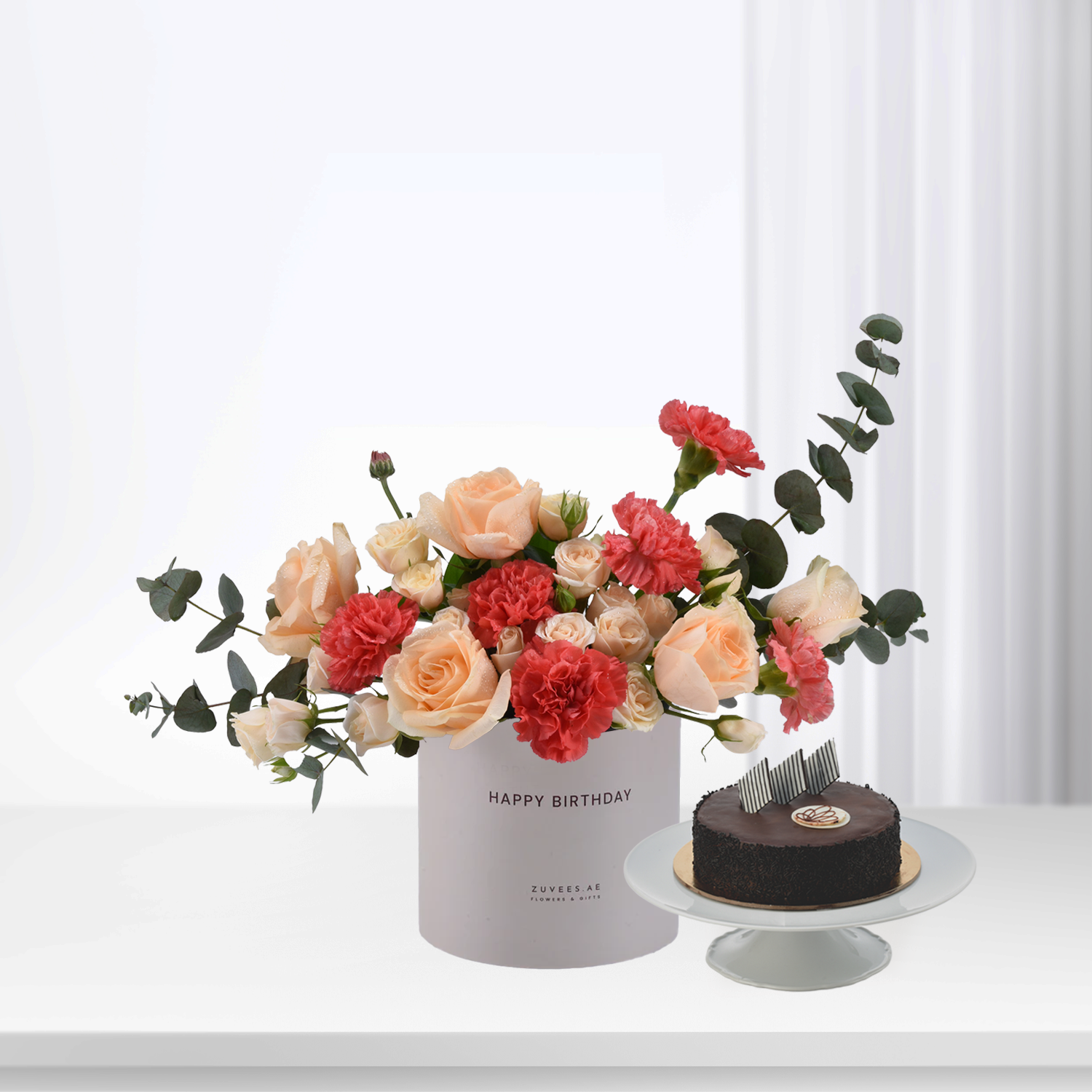 Pastel roses and carnations in white box beside chocolate cake.