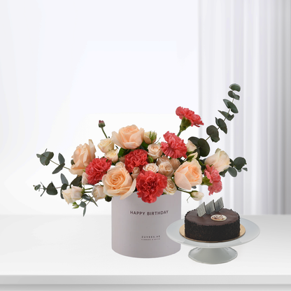 Pastel roses and carnations in white box beside chocolate cake.