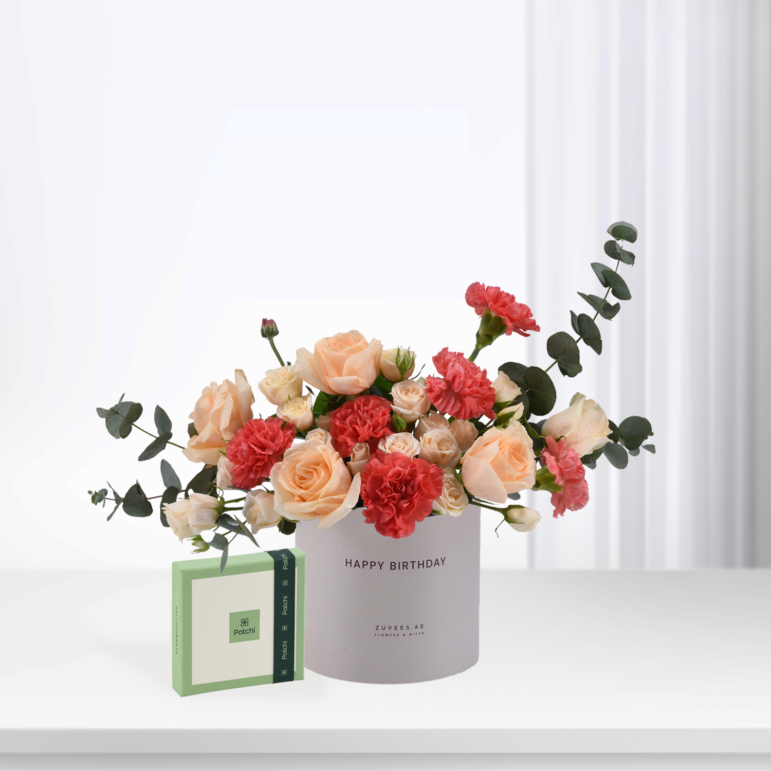 Soft peach roses and red carnations in white "Happy Birthday" vase, beside green Patchi chocolate box.