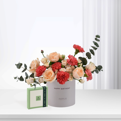 Soft peach roses and red carnations in white "Happy Birthday" vase, beside green Patchi chocolate box.