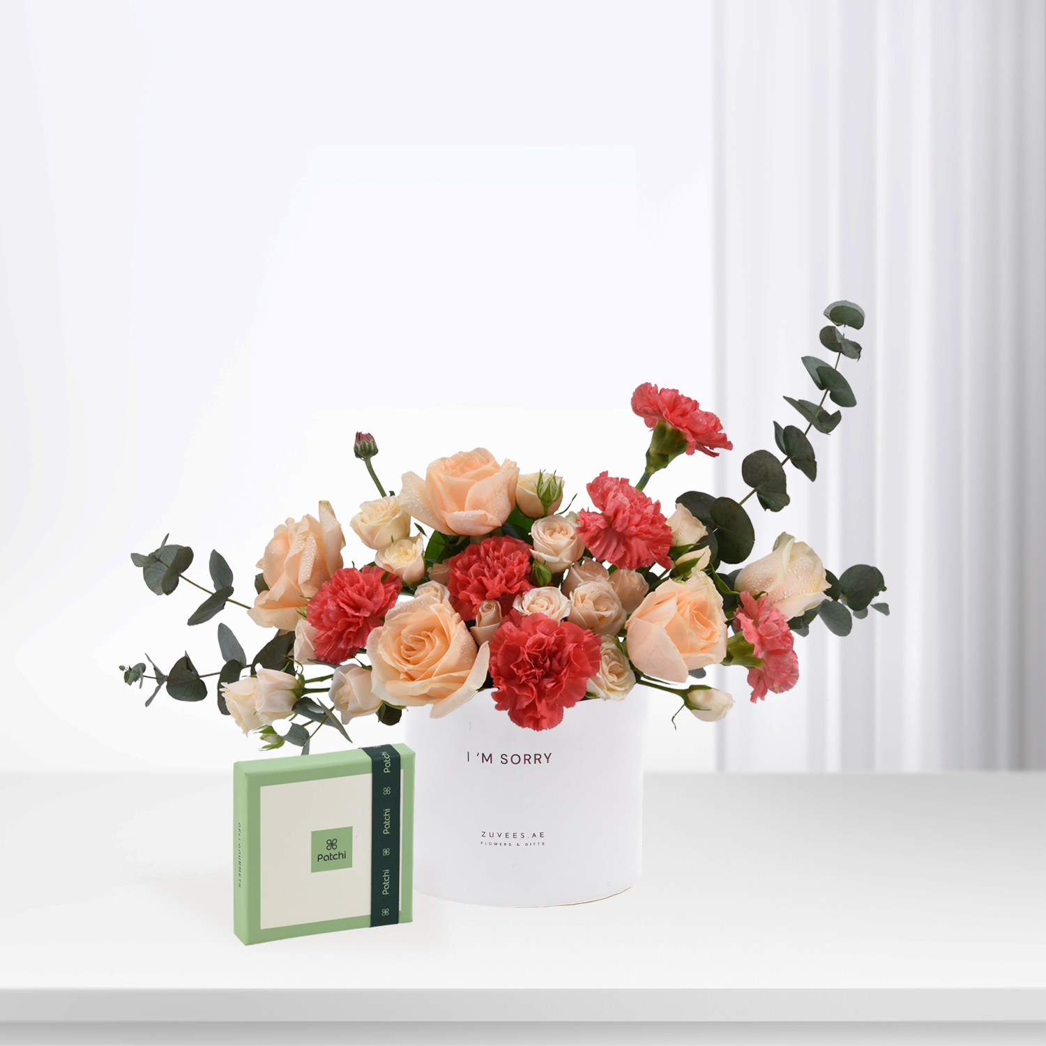 White vase with peach roses and red carnations, beside green Patchi chocolate box.