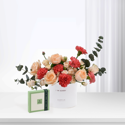 White vase with peach roses and red carnations, beside green Patchi chocolate box.