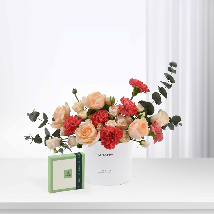 White vase with peach roses and red carnations, beside green Patchi chocolate box.