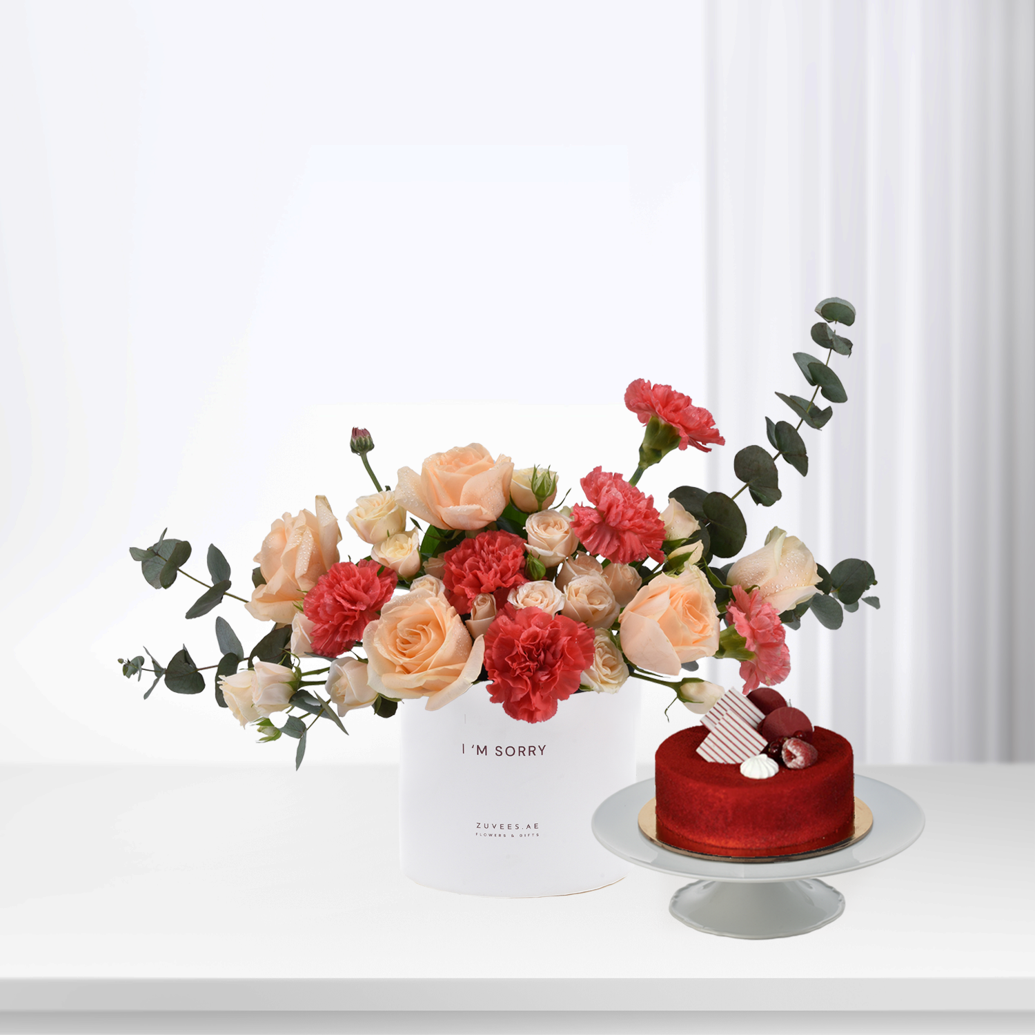 Soft peach roses and red carnations in white box beside red velvet cake.