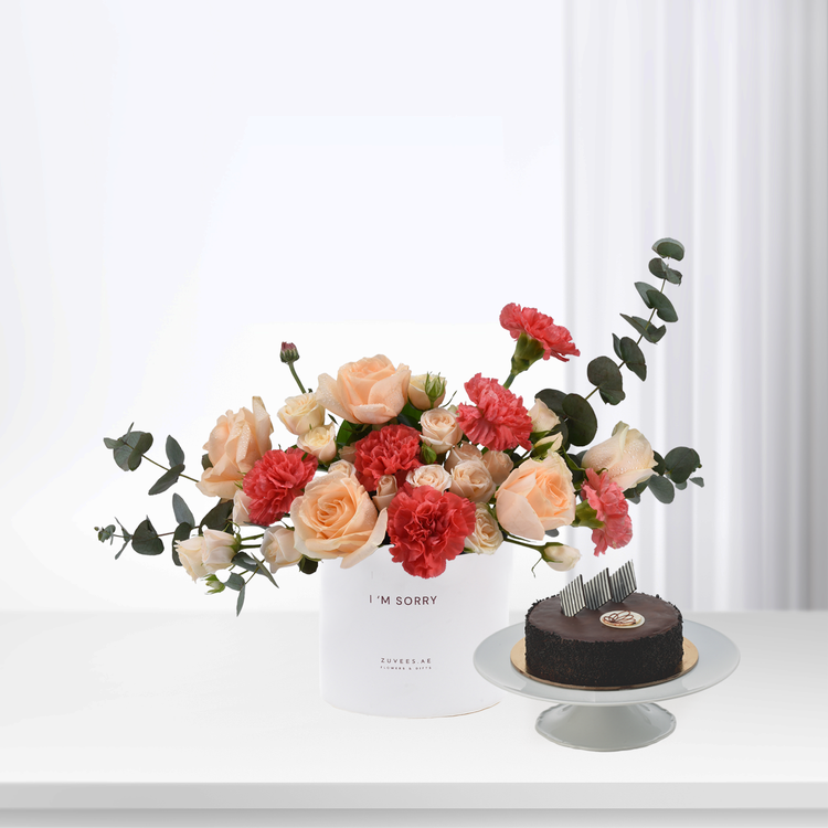 Soft peach roses and red carnations in white box beside chocolate cake.