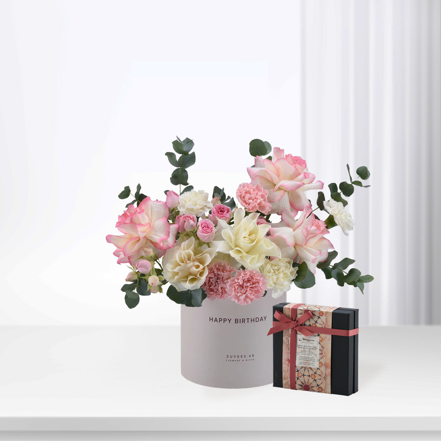 Soft pink and white roses with eucalyptus in branded box, beside decorative gift.