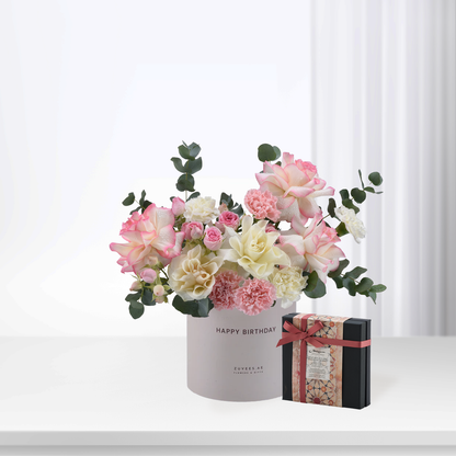 Soft pink and white roses with eucalyptus in branded box, beside decorative gift.