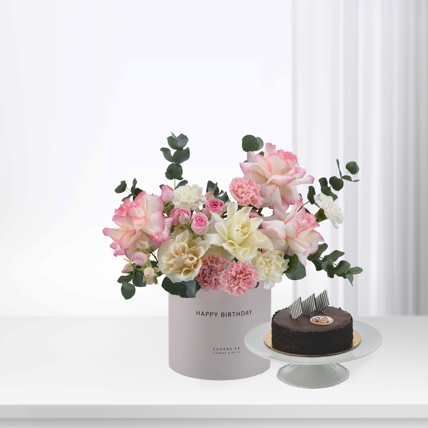 Pink and cream roses, carnations, and eucalyptus in a "Happy Birthday" box beside a chocolate cake.