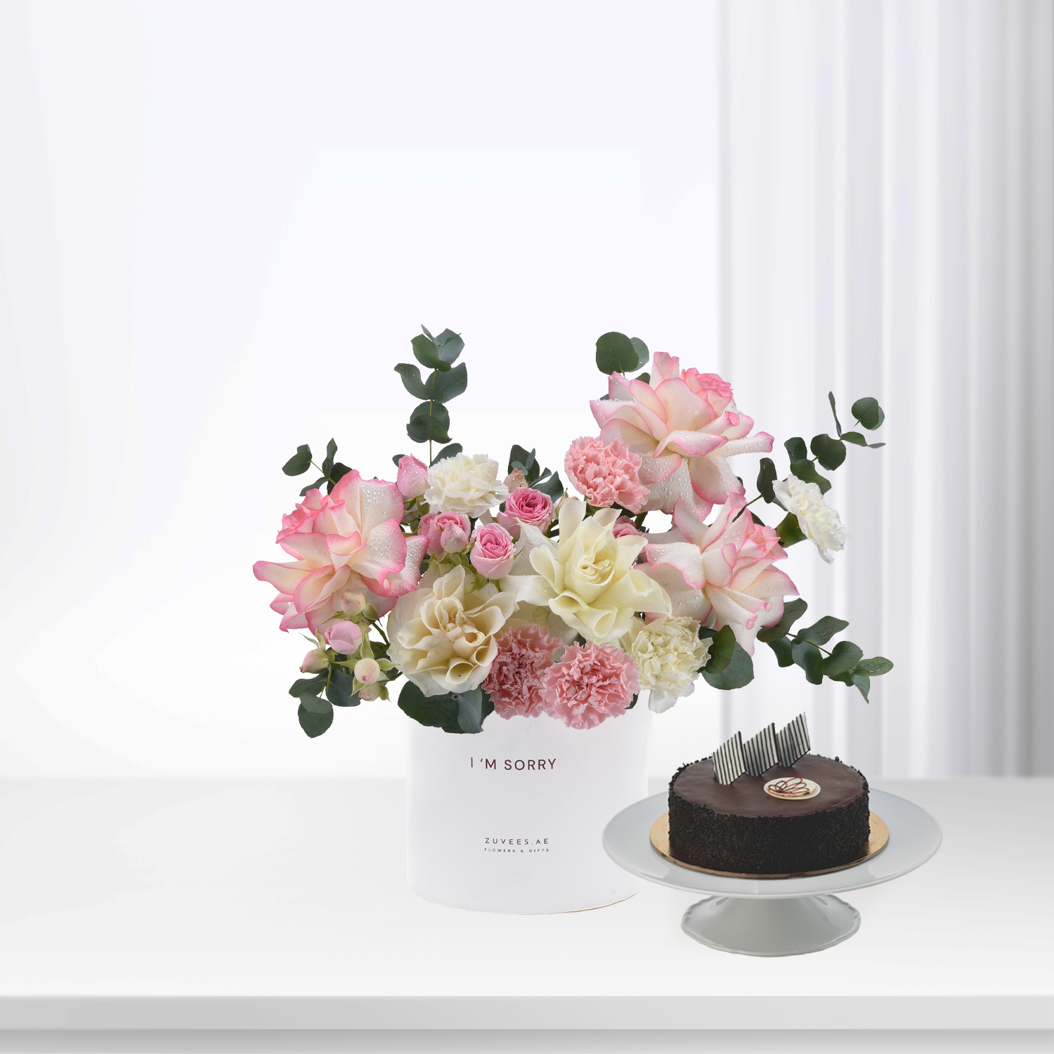 Soft pink roses, carnations, and eucalyptus in white vase beside chocolate cake.