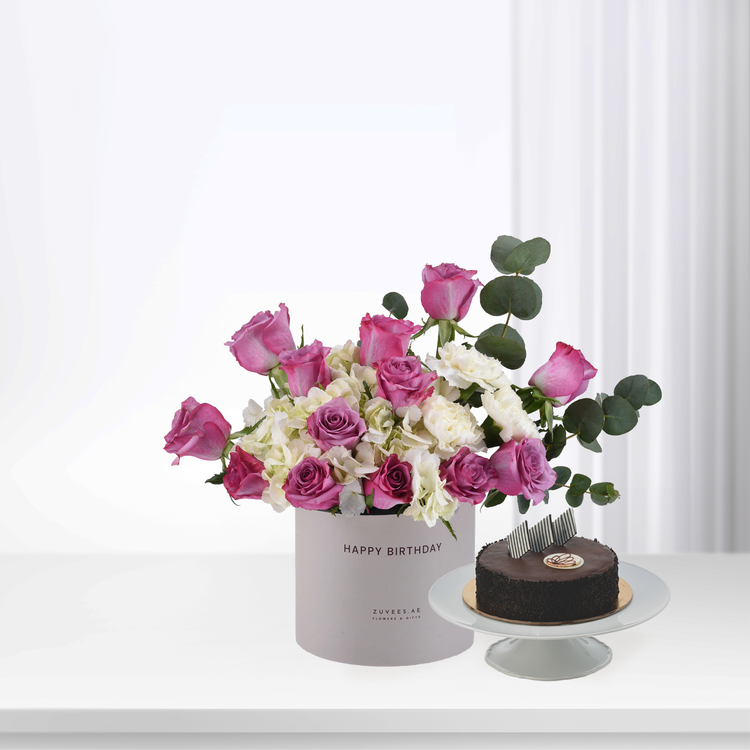 Pink roses, white hydrangeas, and eucalyptus in gray "Happy Birthday" box beside chocolate cake.